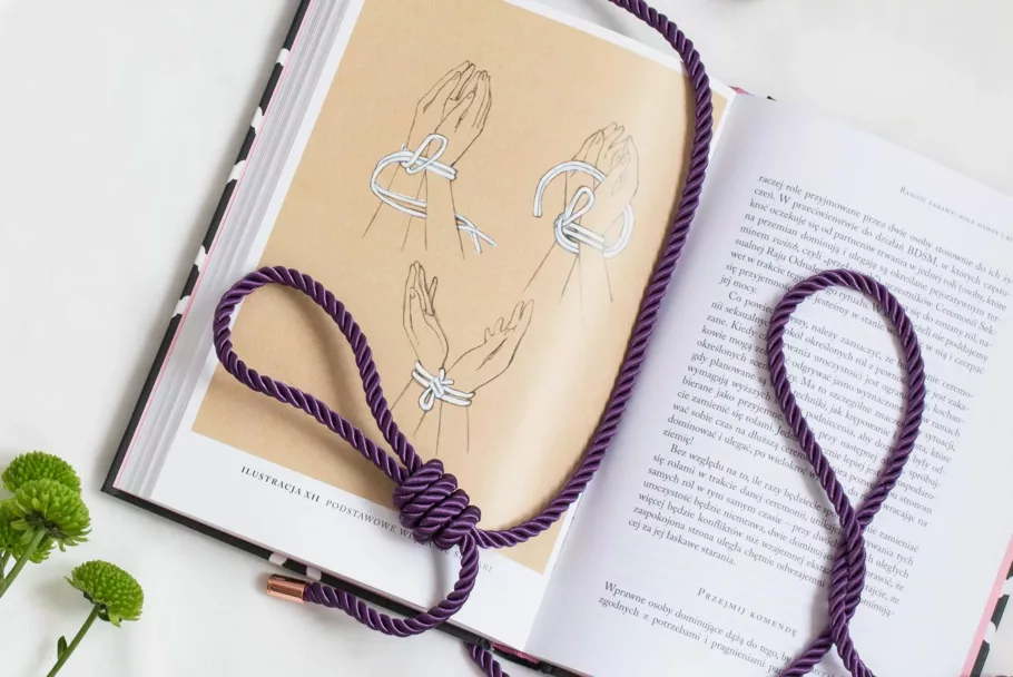 Splątana lina z zawiązaną pętlą leży na książce, otwartej na rysunkowej instrukcji krępowania rąk liną.
