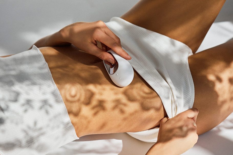 Leżąca osoba, ubrana w krótki biały top i białe szorty trzyma masażer Satisfyer White Temptation nad brzuchem.