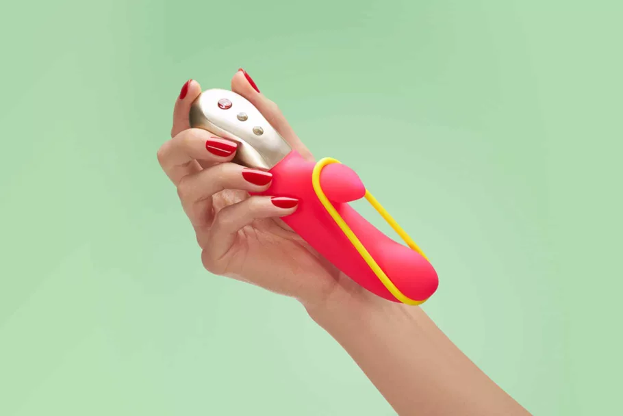 Dłoń trzyma nieduży różowy wibrator króliczek Fun Factory Amorino z żółtą gumką zahaczoną o trzon i mniejszą końcówkę.