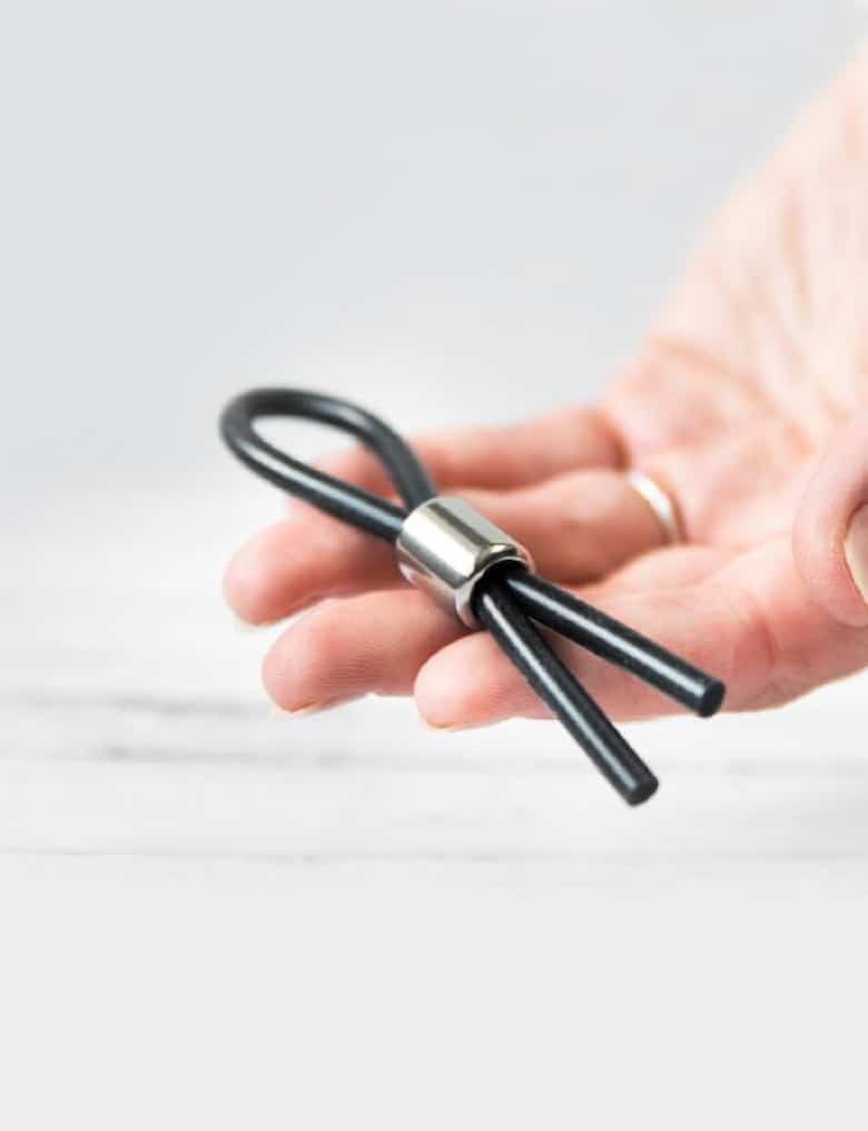 Na dłoni leży lasso erekcyjne Velv'Or JBoa w formie silikonowej linki złożonej na pół i ściśniętej małą, metalową obręczą.
