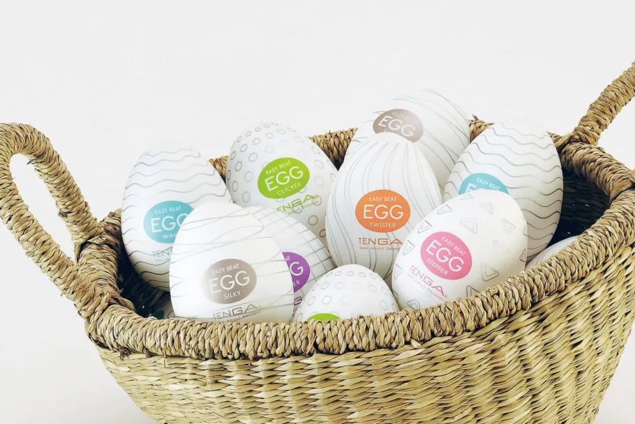 W plecionym koszyku leży 9 białych masturbatorów Tenga Egg w kształcie jajek z kolorowymi logo na etykietach.
