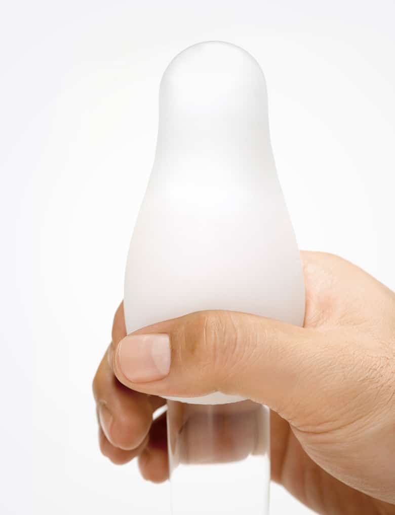 Dłoń obejmuje biały, miękki masturbator Tenga Egg założony na przezroczyste, szklane dildo, rozciągając go wzdłuż gadżetu.