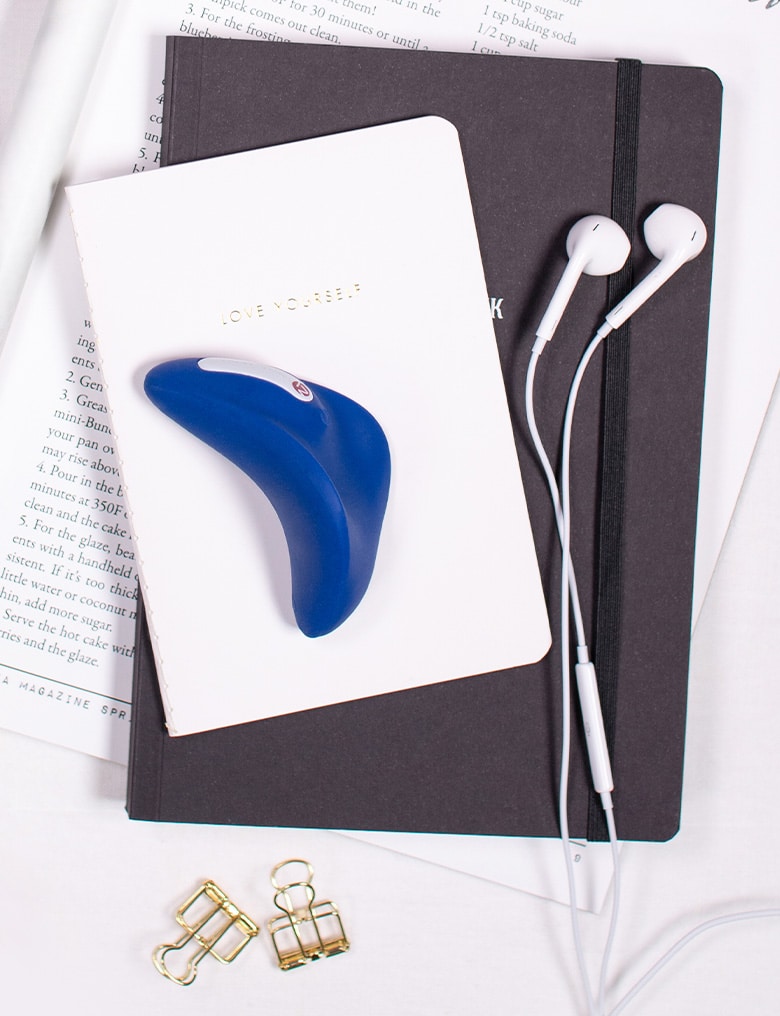 Niebieski masażer Better Than Chocolate 2 leży na notatniku z napisem "Love Yourself". Obok widać douszne słuchawki.