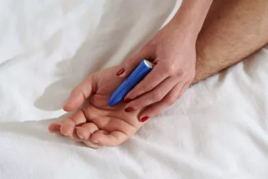 Dłoń z pomalowanymi paznokciami trzyma niebieski masażer w kształcie szminki. Jego czubkiem dotyka wnętrza większej dłoni.