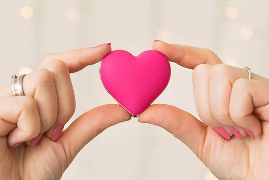 Kciuki i palce wskazujące dwóch dłoni trzymają różowy masażer łechtaczki Rianne S Heart Vibe w kształcie serca.