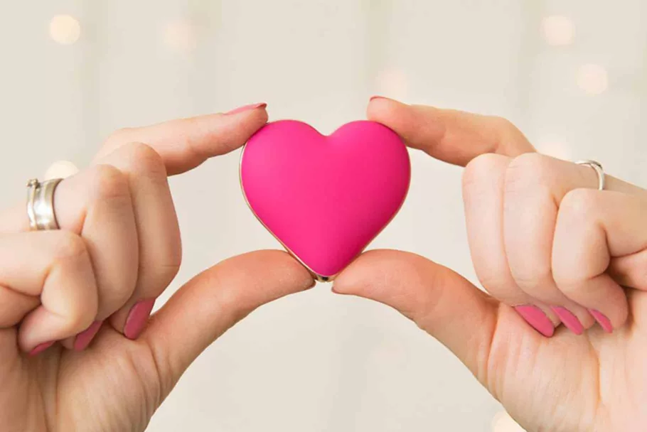 Kciuki i palce wskazujące dwóch dłoni trzymają różowy masażer łechtaczki Rianne S Heart Vibe w kształcie serca.