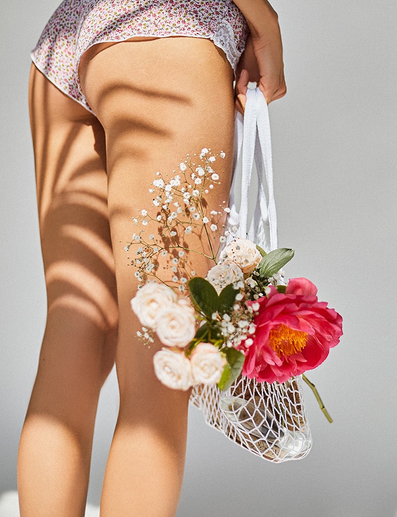 Modelka ubrana w same majtki we wzór drobnych, różowych kwiatków trzyma białą torbę, z której wystaje bukiet kwiatów.