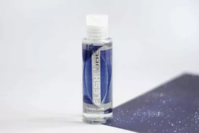 Przezroczysta buteleczka lubrykantu FleshLube Water z granatową etykietą stoi na kartce z ilustracją gwieździstego nieba.