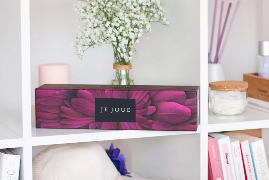 Opakowanie wibratora Je Joue leży na półce. Powierzchnię kartonika zdobi ilustracja ciemnoróżowych piór oraz logo marki.