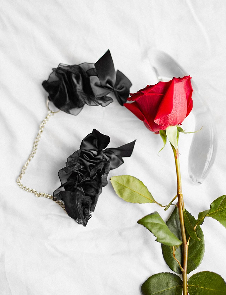 Na pościeli leży róża i kajdanki Bijoux Indiscrets Frou Frou w formie bransoletek z czarnej organzy połączonych łańcuszkiem.