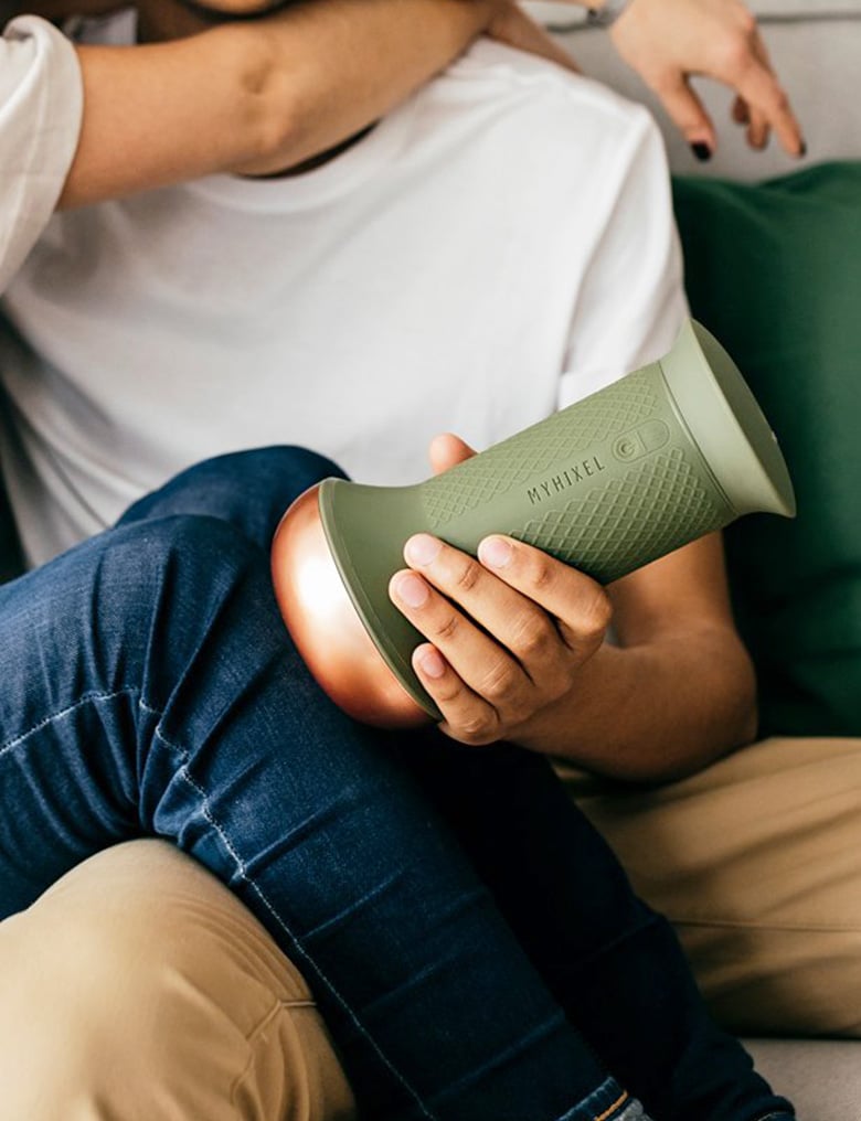 Osoba ubrana w t-shirt i dżinsy siedzi na kolanie partnera. On trzyma przy jej kolanie zielono-miedziany masturbator MyHixel.