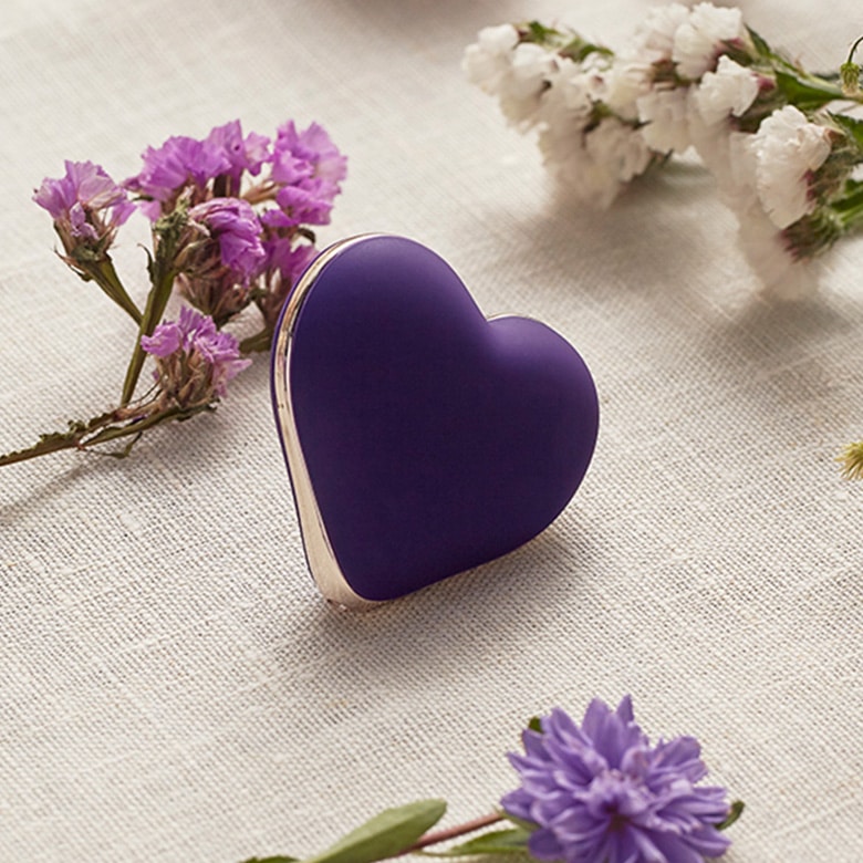 Fioletowy, matowy masażer-serduszko Rianne S Heart Vibe z silikonu leży na lnianym obrusie. Wokół niego widać drobne kwiaty.