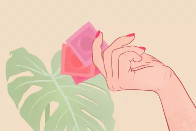 Dłoń z pomalowanymi paznokciami trzyma trzy prezerwatywy w różowych i czerwonych foliach. W tle widać liść monstery.