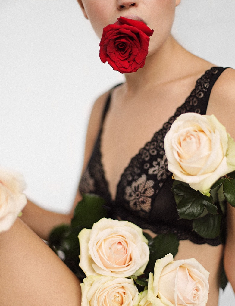 Osoba w czarnej bieliźnie siedzi na podłodze. W ustach ma kwiat czerwonej róży, a na udach i pod pachą trzyma jasne róże.