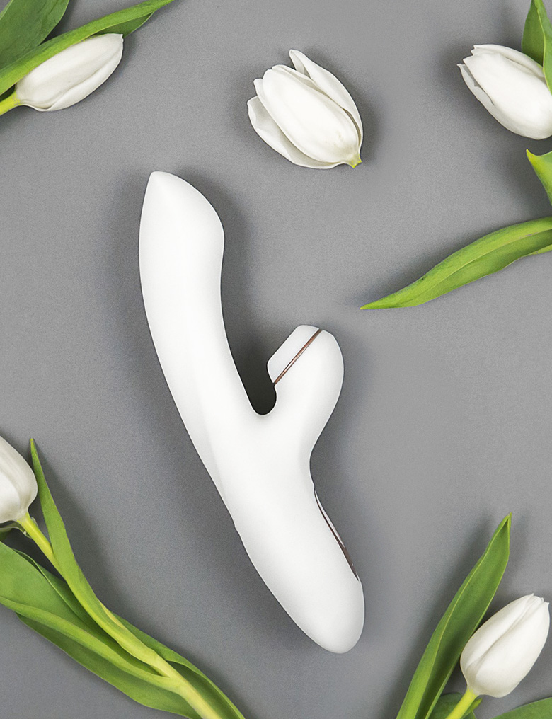 Biały króliczek Satisfyer Pro + G-Spot z bezdotykową końcówką do stymulacji łechtaczki leży na szarym tle wśród tulipanów.