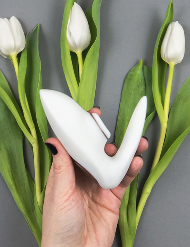 Dłoń trzyma wibrator dla par Satisfyer Pro 4 Couples z bezdotykową częścią. Pod nimi na szarym tle leżą trzy białe tulipany.