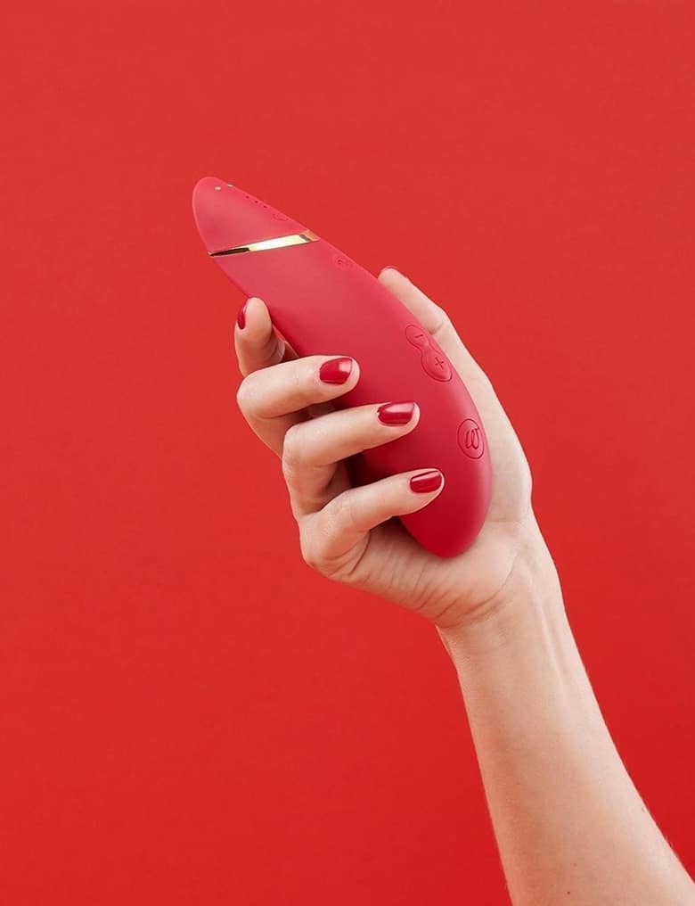 Na czerwonym tle dłoń z pomalowanymi na bordowo paznokciami trzyma duży, czerwony masażer bezdotykowy Womanizer Premium 2.