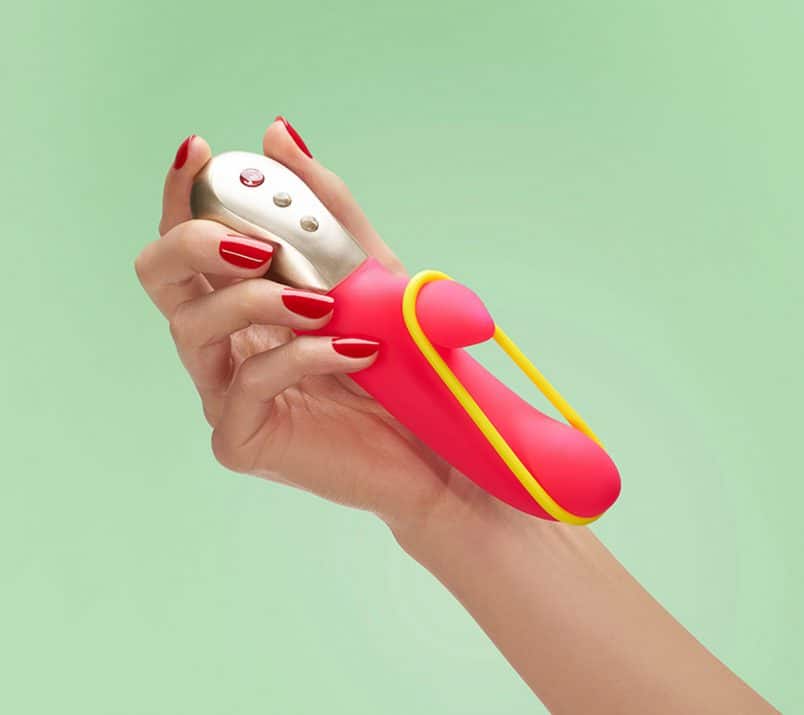 Dłoń trzyma nieduży różowy wibrator króliczek Fun Factory Amorino z żółtą gumką zahaczoną o trzon i mniejszą końcówkę.