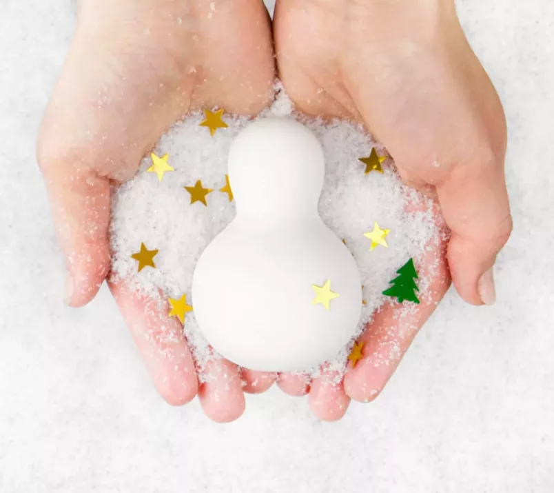 Masażer-bałwanek Iroha Yuki leży w dłoniach posypanych śniegiem i złotym konfetti w kształcie gwiazdek.