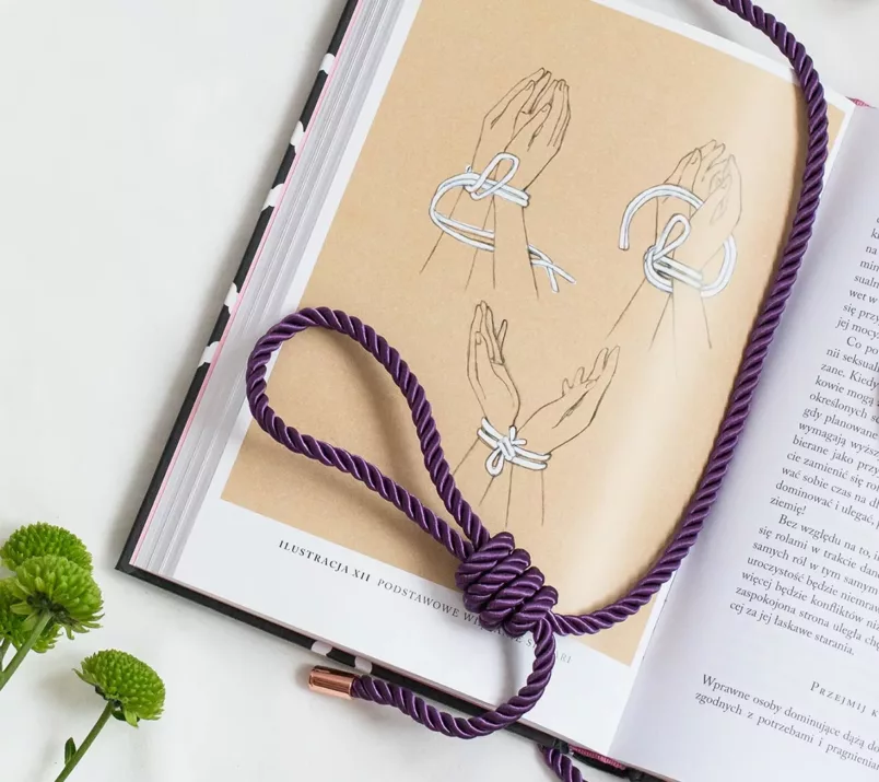 Splątana lina z zawiązaną pętlą leży na książce, otwartej na rysunkowej instrukcji krępowania rąk liną.