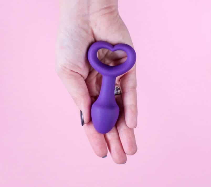 Na wyciągniętej dłoni leży mały, fioletowy korek analny Lovelife Explore z uchwytem w kształcie serduszka.