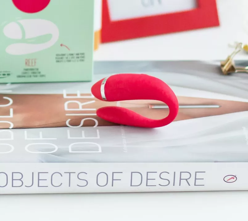 Na książce o tytule "Objects of desire" leży czerwony wibrator dla par Moqqa Reef by We-Vibe i jego opakowanie.