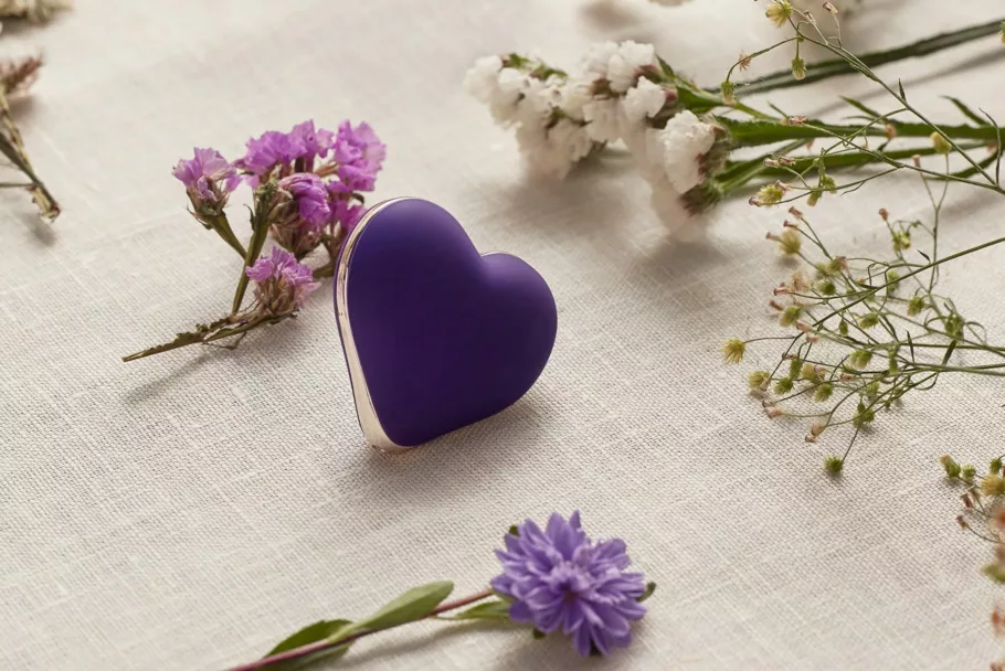Fioletowy masażer-serduszko Rianne S Heart Vibe leży na lnianej tkaninie. Wokół niego widać drobne, fioletowe i białe kwiaty.