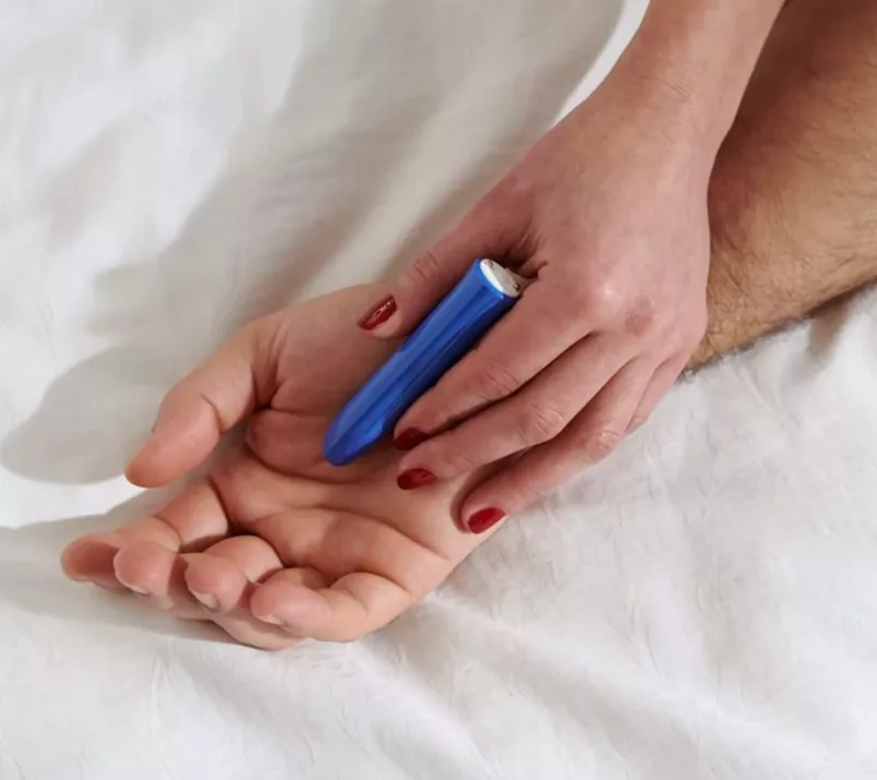 Dłoń z pomalowanymi paznokciami trzyma niebieski masażer w kształcie szminki. Jego czubkiem dotyka wnętrza większej dłoni.