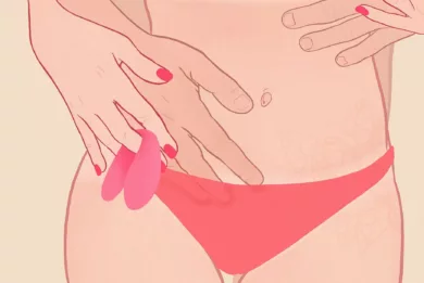 Osoba z czerwonymi paznokciami przy biodrze trzyma na palcu różowy wibrator dla par. Partner obejmuje ją rękami od tyłu.