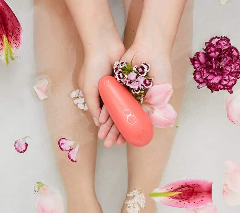 Womanizer Starlet 2 wraz z kwiatami leży w dłoniach osoby w wannie. W tle widać łydki osoby i kwiaty pływające w wodzie.