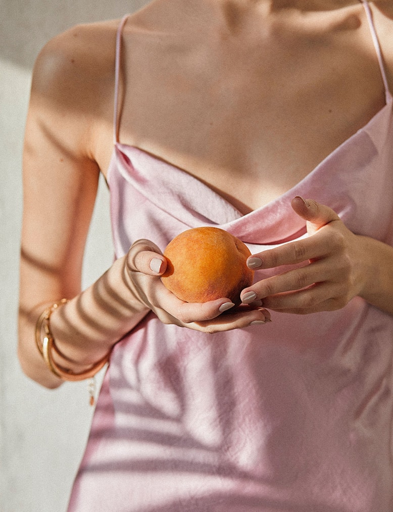Postać ubrana w jasnoróżową sukienkę, ze złotą bransoletą na ręce przekłada brzoskwinię z jednej dłoni do drugiej.