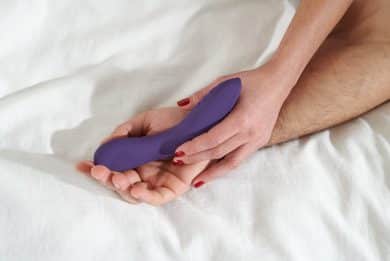 Dłoń z pomalowanymi paznokciami trzyma fioletowy, wygięty wibrator. Jego czubkiem dotyka wnętrza większej dłoni.