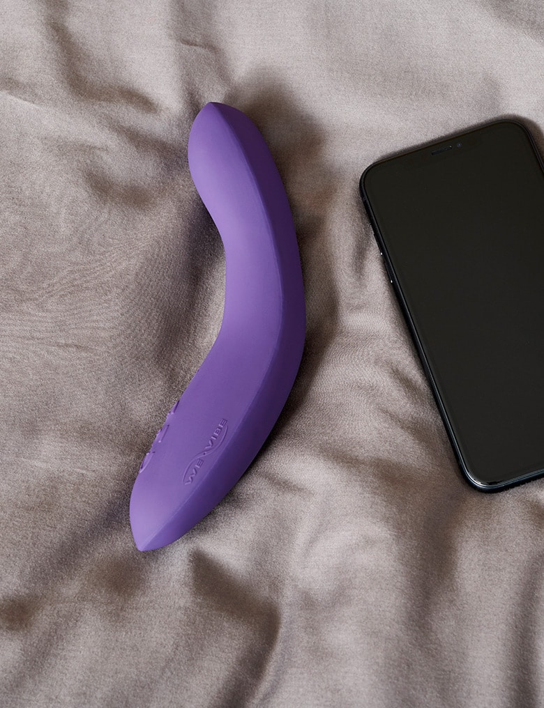 Na szarym pledzie obok smartfona leży fioletowy, wygięty wibrator We-Vibe Rave z opcją sterowania aplikacją.