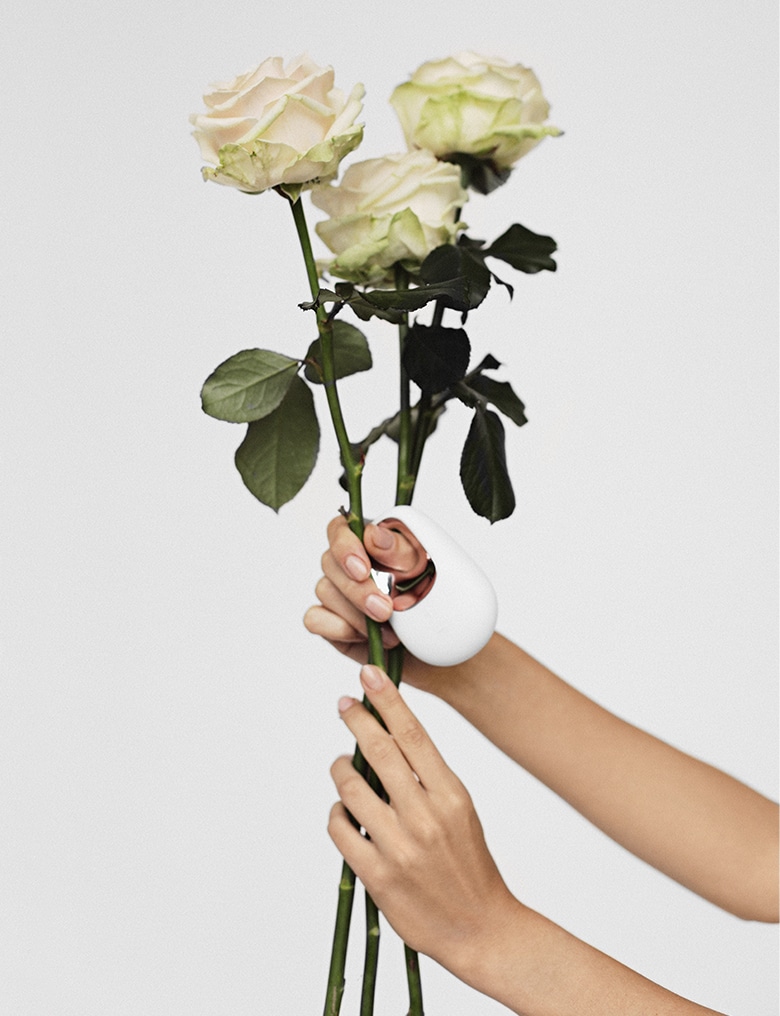 Dłonie trzymają trzy białe róże. Na górnym kciuku zawieszony jest biało-złoty masażer White Temptation.