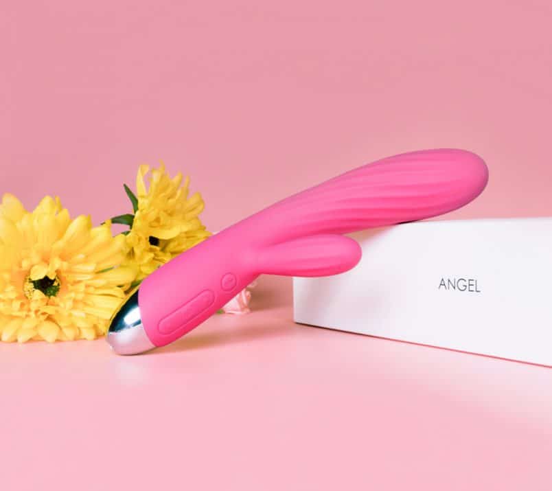 Intensywnie różowy wibrator króliczek leży oparty o biały kartonik z napisem "Angel". W tle leżą duże, żółte kwiaty.