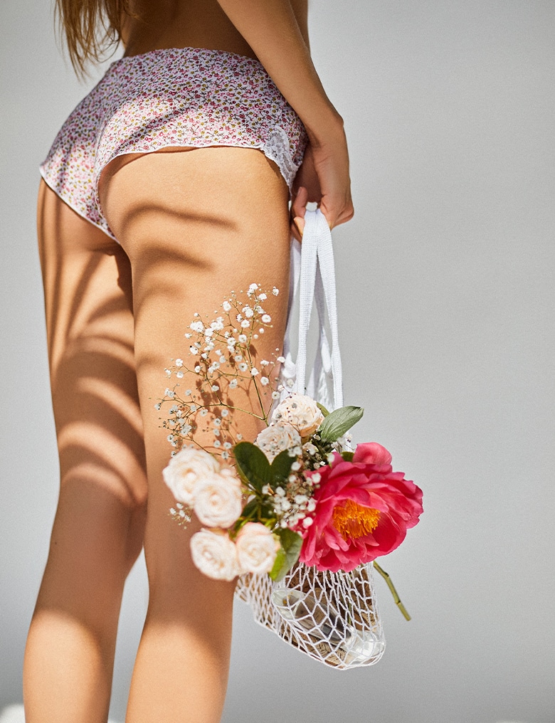 Widoczna od tyłu modelka ubrana w same majtki trzyma przy nodze plecioną, białą siatkę, z której wystaje bukiet kwiatów.