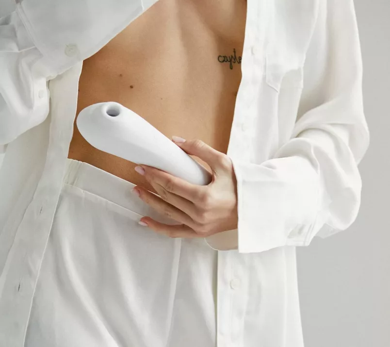 Osoba ubrana w białe spodnie i rozpiętą koszulę trzyma biały masażer Satisfyer Dual Pleasure przy brzuchu.