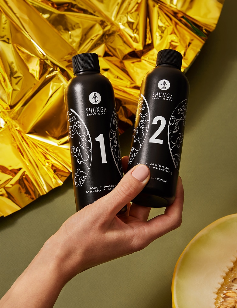 Dłoń trzyma czarne, minimalistyczne butelki zestawu Shunga do erotycznego masażu. W tle widać złote prześcieradło.