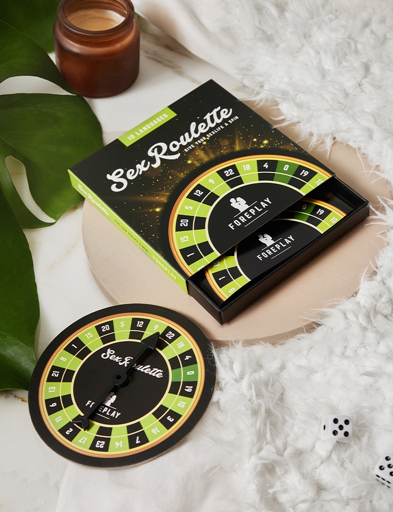 Opakowanie i tarcza gry Sex Roulette w zielonej wersji Foreplay leżą na jasnym, ozdobnym tle obok liścia monstery.