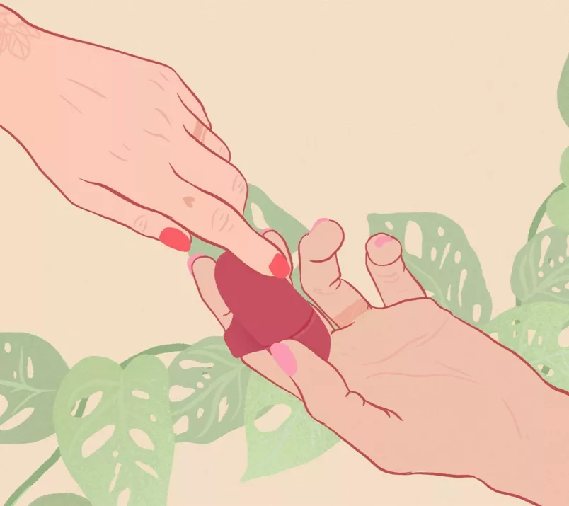 Na rysunku masażer łechtaczki jest założony na palec jednej dłoni. Druga dłoń dotyka powierzchni gadżetu. W tle widać liście.