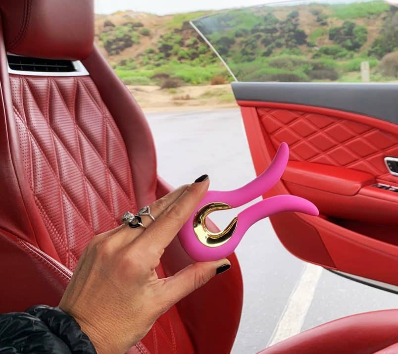 Dłoń trzyma różowy wibrator Gvibe MINI w samochodzie z czerwoną tapicerką. Za otwartymi drzwiami widać zielone pagórki.