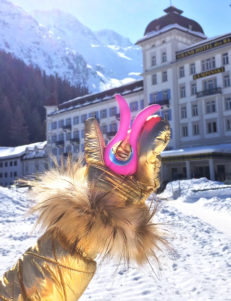 Różowy wibrator Gvibe MINI trzymany przez dłoń w złotej, zimowej rękawicy. W tle widać hotel w wysokich, ośnieżonych górach.