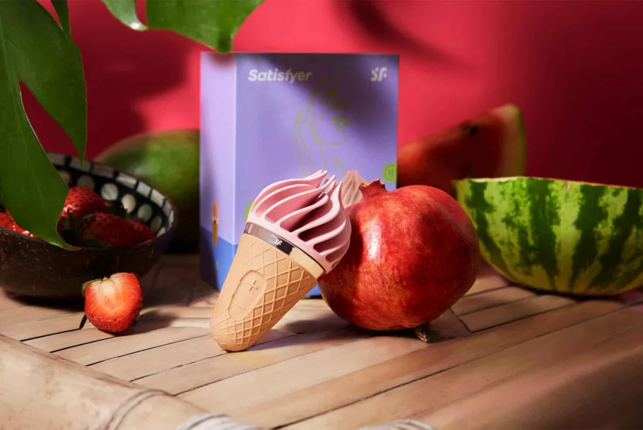 Masażer w kształcie loda włoskiego oparty jest o owoc granatu. Na czerwonym tle widać fioletowe opakowanie i więcej owoców.