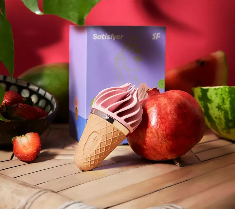 Masażer w kształcie loda włoskiego oparty jest o owoc granatu. Na czerwonym tle widać fioletowe opakowanie i więcej owoców.