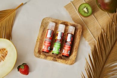 Trzy buteleczki lubrykantów System JO Tri Me Flavors leżą na małej drewnianej tacce. Wokół nich widać owoce i beżowe ozdoby.