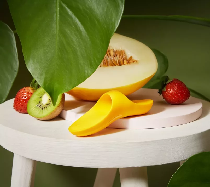 Pod dużym liściem na drewnianym stoliku leży żółty masażer Satisfyer Teaser, otoczony truskawkami, połówką kiwi i melona.
