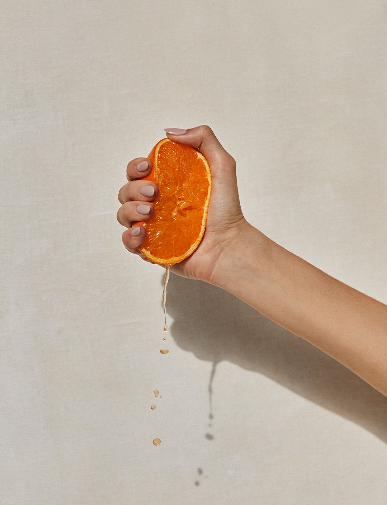 Na gładkim, jasnoszarym tle dłoń ściska mocno połówkę pomarańczy, z której ciekną krople soku.