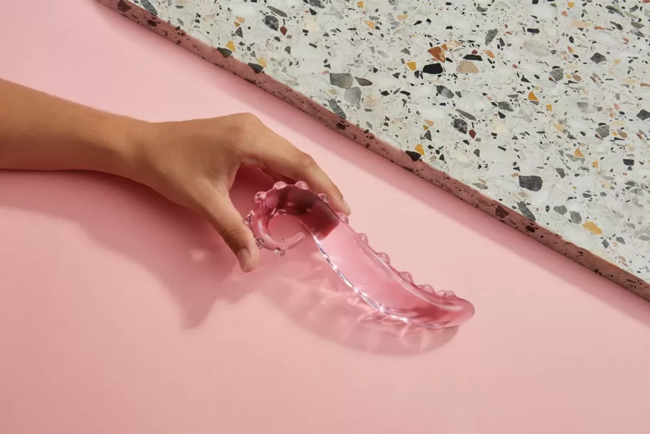 Dłoń trzyma szklane dildo Gläs Lick It na ozdobnym tle: częściowo różowym, częściowo złożonym z lastryko.