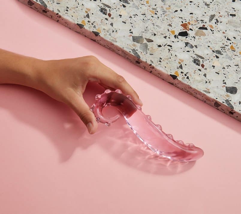 Dłoń trzyma szklane dildo Gläs Lick It na ozdobnym tle: częściowo różowym, częściowo złożonym z lastryko.
