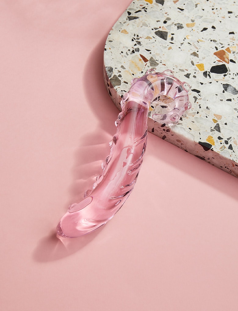 Różowe, szklane dildo Gläs Lick It leży na różowym tle oparte o płytę lastryko.